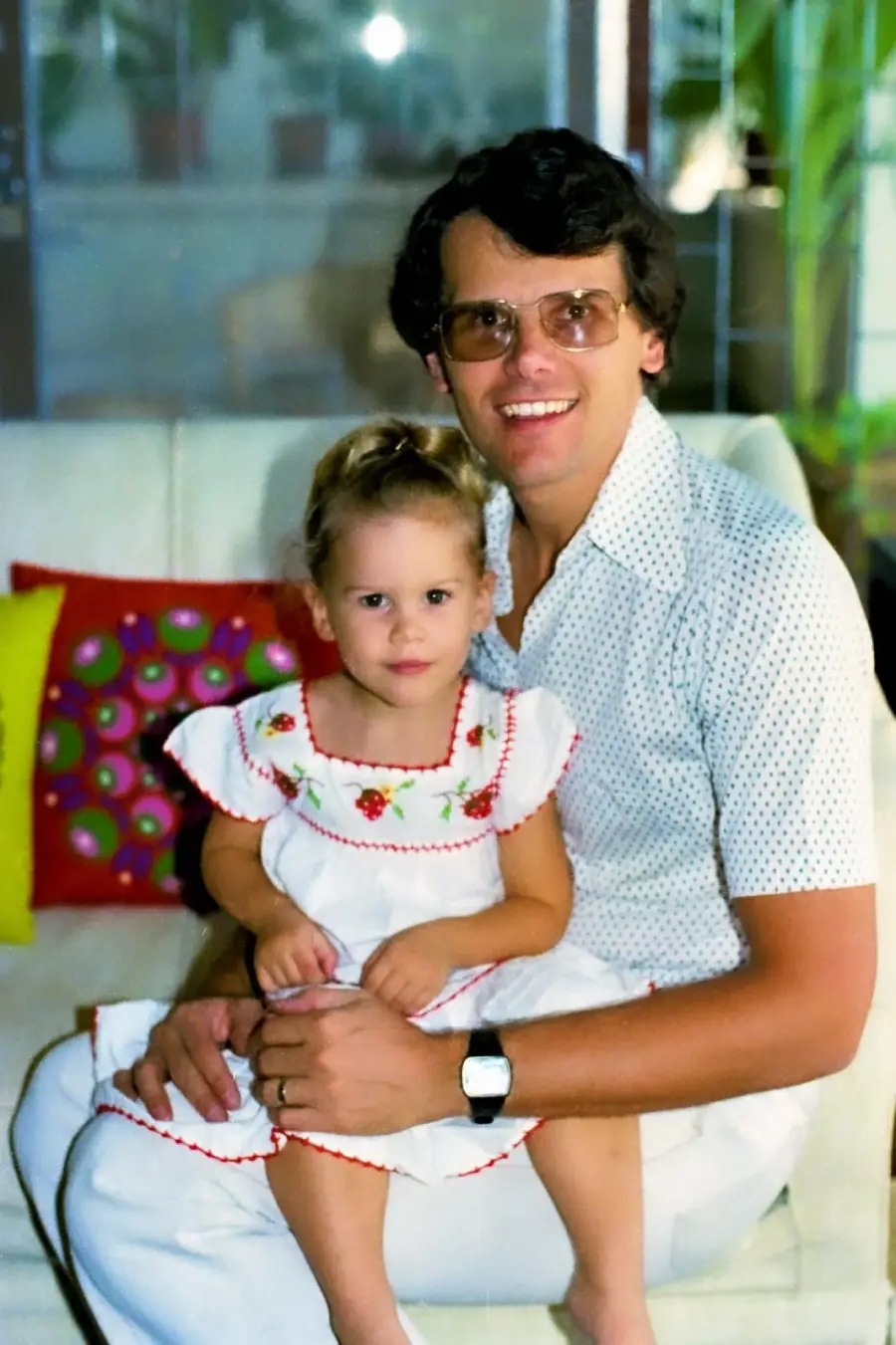 A man in glasses and a white shirt sits on a couch, smiling, with a young girl in a white dress with red trim and floral embroidery on his lap. There are colorful pillows and green plants in the background.