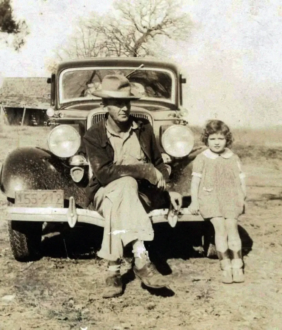 An older man in a hat and a young girl stand in front of a vintage car on a dirt road, with bare trees and a wooden house in the background; the photo appears old and sepia-toned.