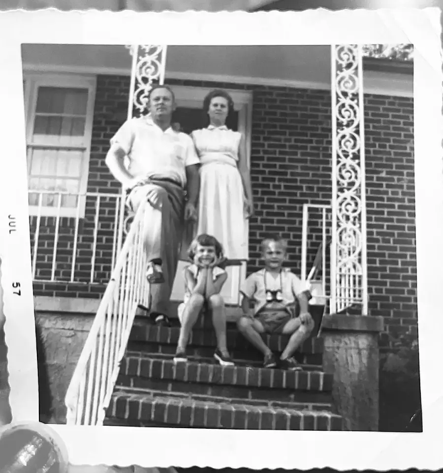 A black-and-white photo of a man and woman standing on a brick porch with decorative railings, and two young children sitting on the steps in front of them, taken in July 1957.