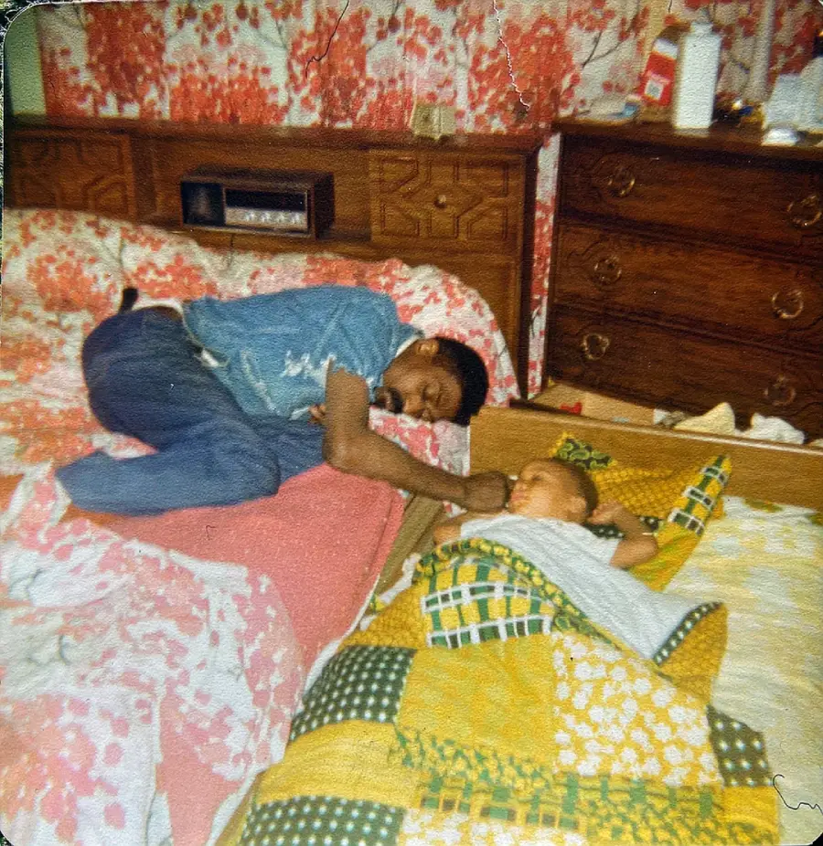 A man and a baby are sleeping on separate beds in a bedroom with colorful, floral wallpaper. The man rests on a pink bed, while the baby lies on a patchwork quilt. A wooden dresser with various items is in the background.