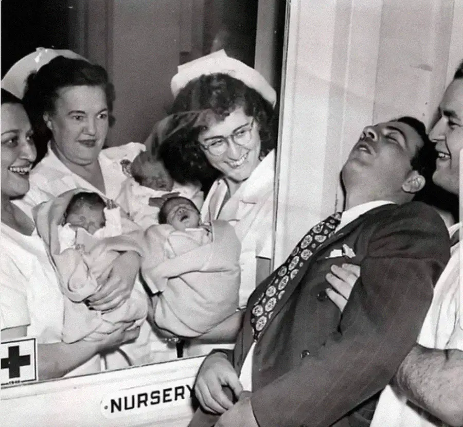 Three nurses stand behind a window holding newborn babies and smiling. In front of the window, a man in a suit appears to have fainted while another man supports him. The word "NURSERY" is visible on the window.