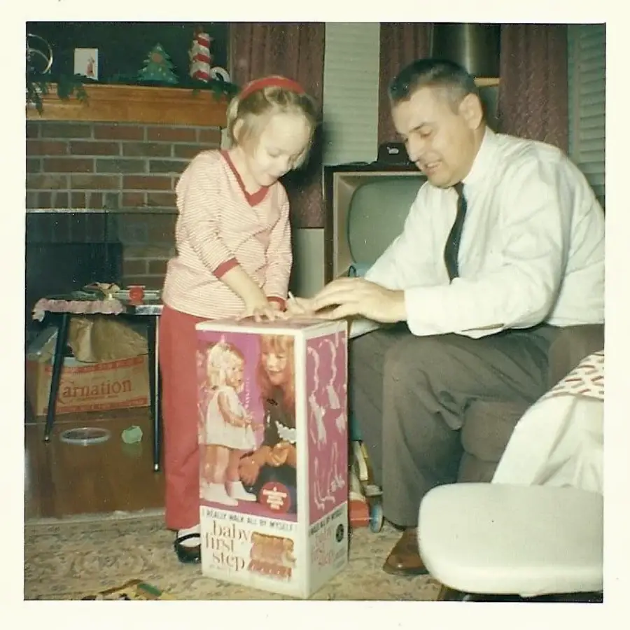 A young girl opens a "Baby First Step" doll box with the help of a man, likely her father, in a vintage living room with a brick fireplace and a television in the background.