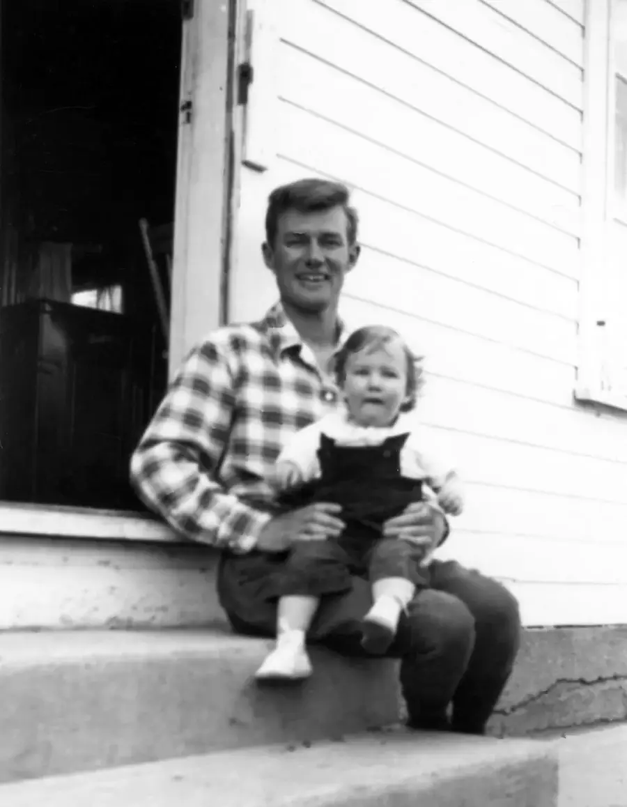 A man in a plaid shirt sits on outdoor steps with a young child on his lap, in front of a light-colored wooden building with an open door. The photo is black and white.