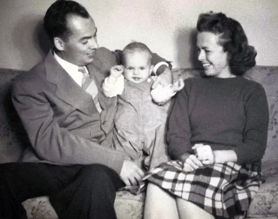 A black-and-white photo of a man in a suit, a woman in a plaid skirt, and a smiling baby sitting together on a couch, all looking happy and engaged.