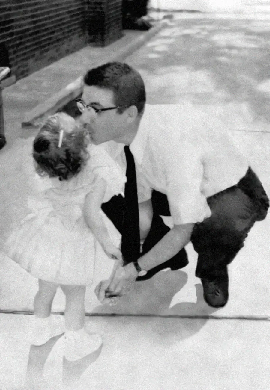 A man in glasses kneels on a sidewalk to kiss a small girl who is dressed in a light-colored dress and shoes. The scene appears to be from the past, in black and white.