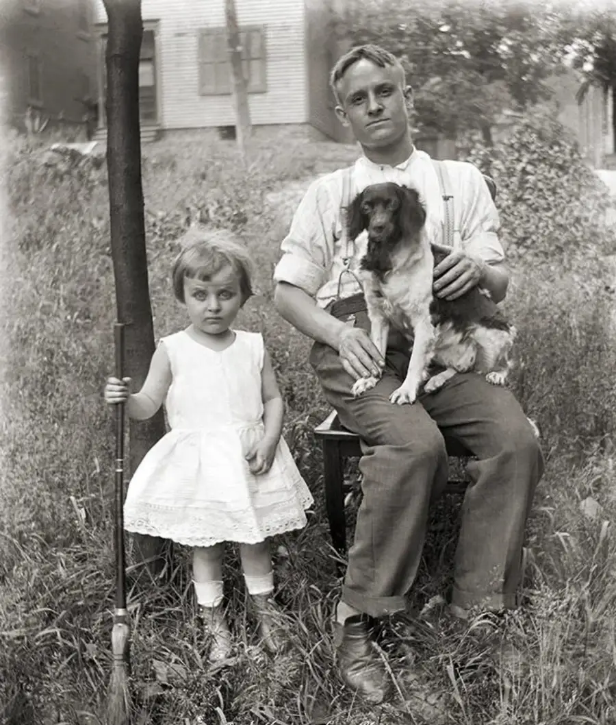 A man sits on a bench holding a dog, while a young girl in a white dress stands beside him holding a rifle. They are outdoors in a grassy yard with a house and trees in the background.