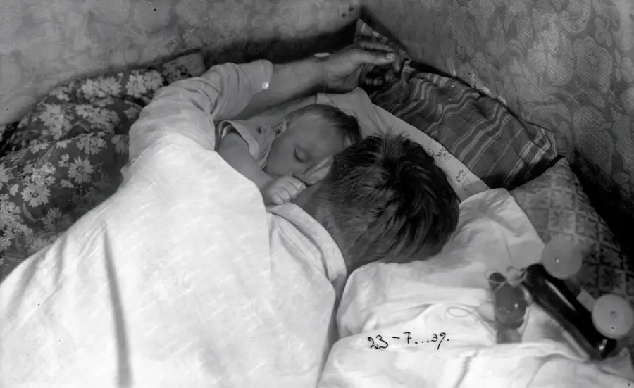 A man lies on a bed, hugging a sleeping baby close to his chest. Both are resting on patterned pillows and blankets. A pair of baby bottles lies nearby, and a date, 23-7-39, is written on the photo.