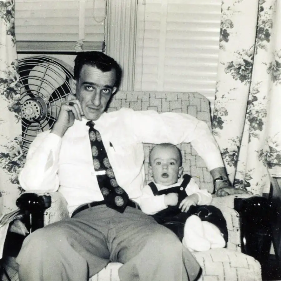 A man in a white shirt and patterned tie sits in an armchair with a baby on his lap. The baby wears a light shirt and dark overalls. They are in a room with floral curtains and a fan in the background.