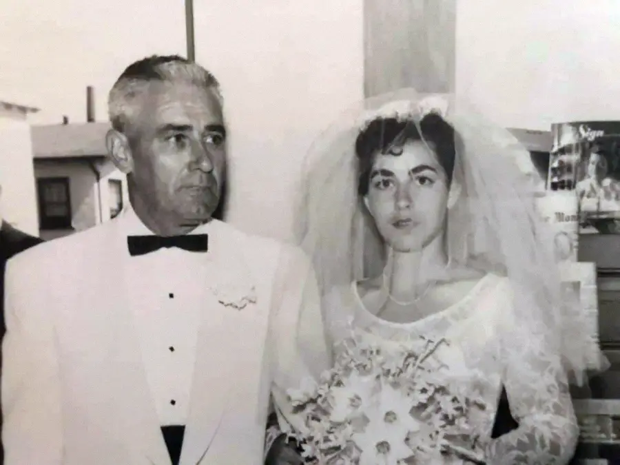 A black and white photo of a bride in a wedding dress and veil holding a bouquet, standing next to an older man in a white tuxedo jacket with a bow tie, likely at a wedding ceremony.