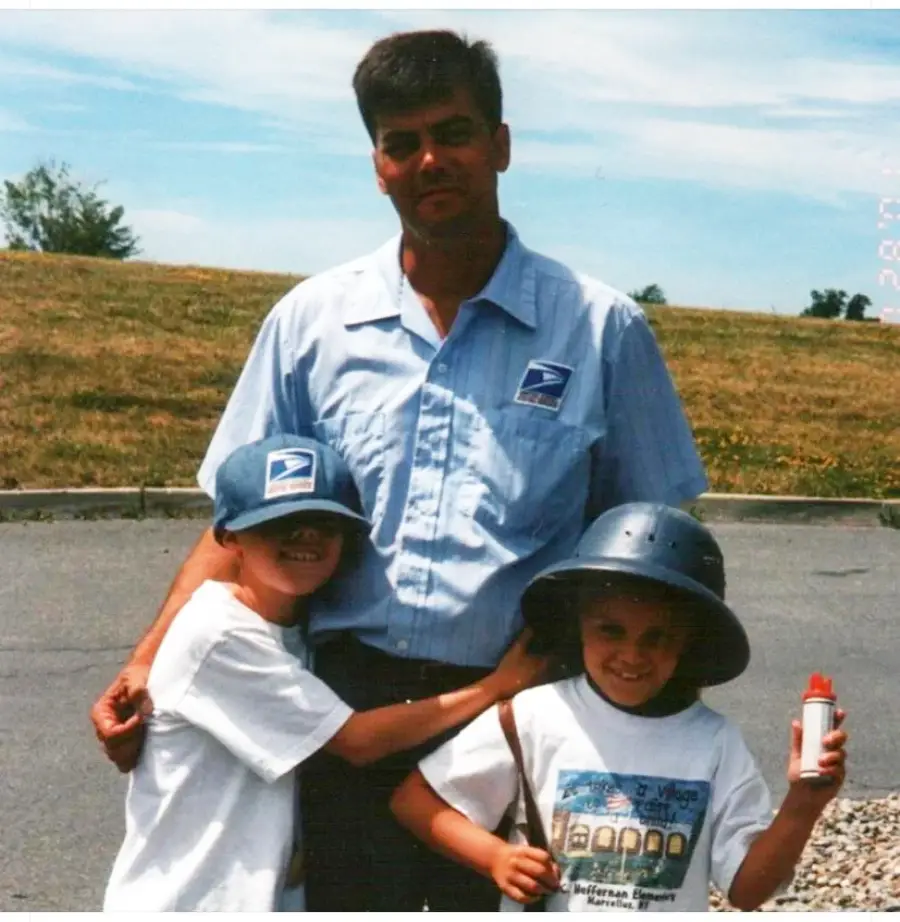 A man in a USPS uniform stands outside with two smiling children, one hugging him and wearing a USPS cap, the other wearing a helmet and holding a can. They are on a paved area near grass under a blue sky.