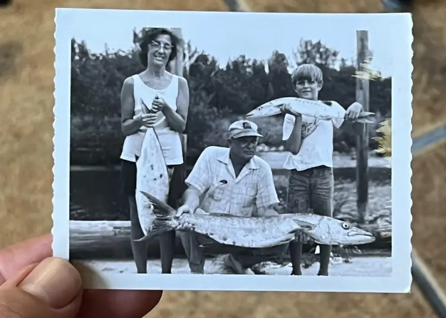 A hand holds a vintage photo showing a smiling woman, a seated man, and a boy, all proudly displaying large fish they caught outdoors, with trees in the background.