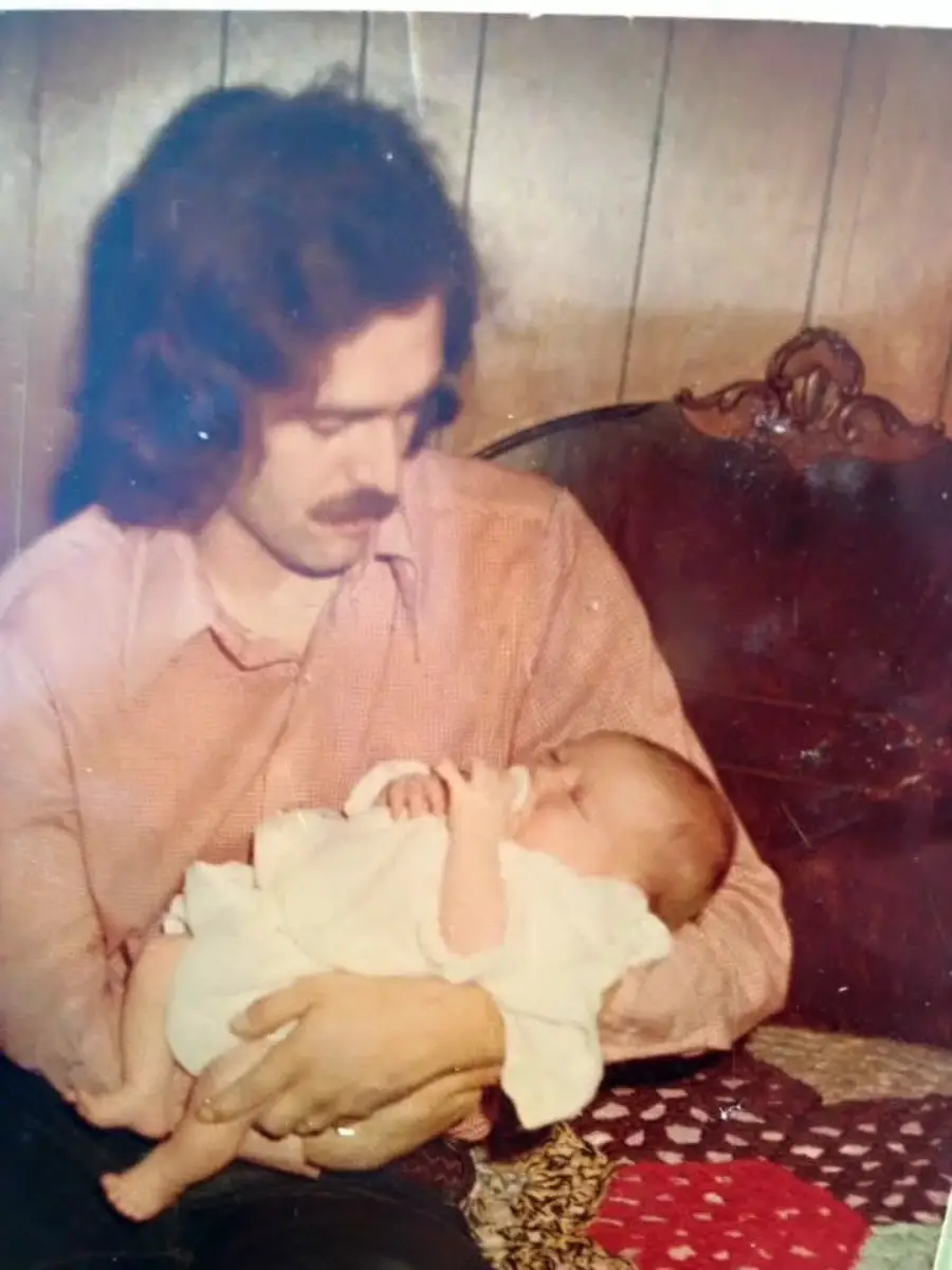 A man with wavy hair and a mustache sits on a patterned quilt, feeding a baby with a bottle. The baby is wrapped in a light-colored blanket, and the background features wood-paneled walls and a carved headboard.
