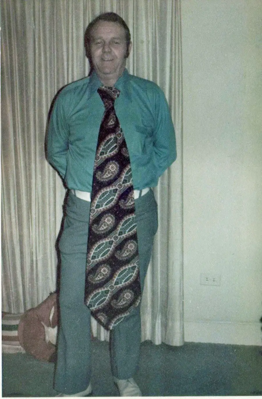A man stands indoors with his hands behind his back, wearing a teal shirt, blue pants, and a very large, patterned necktie that extends down past his knees. Beige curtains and part of a wall are visible behind him.