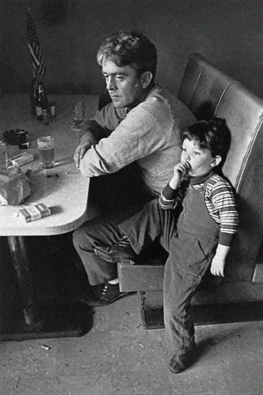 A man sits at a table in a booth, looking away, with a drink and cigarettes on the table. A young child in overalls stands beside him, leaning against the man and sucking their thumb.