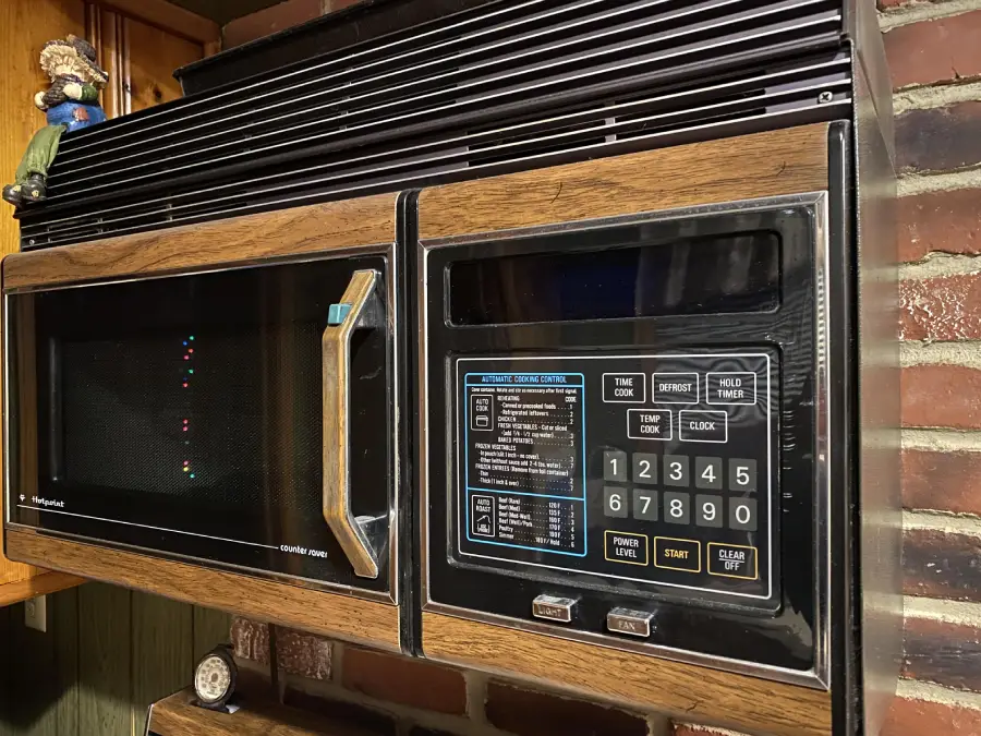 A vintage microwave oven with a woodgrain finish, digital keypad, and display sits against a brick wall. The control panel has various buttons and instructions, and a small figurine is visible on the shelf above.