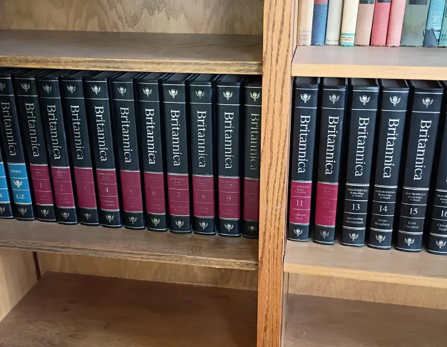 A wooden bookshelf holds volumes of the Encyclopaedia Britannica, with black and red spines labeled with volume numbers and titles. Some other books are visible on the shelf above.