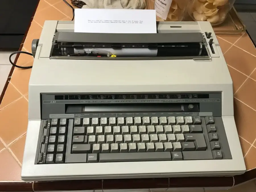 A beige electronic typewriter with a gray keyboard sits on a tiled surface. A sheet of paper with text is inserted in the typewriter. Various objects are partially visible in the background.