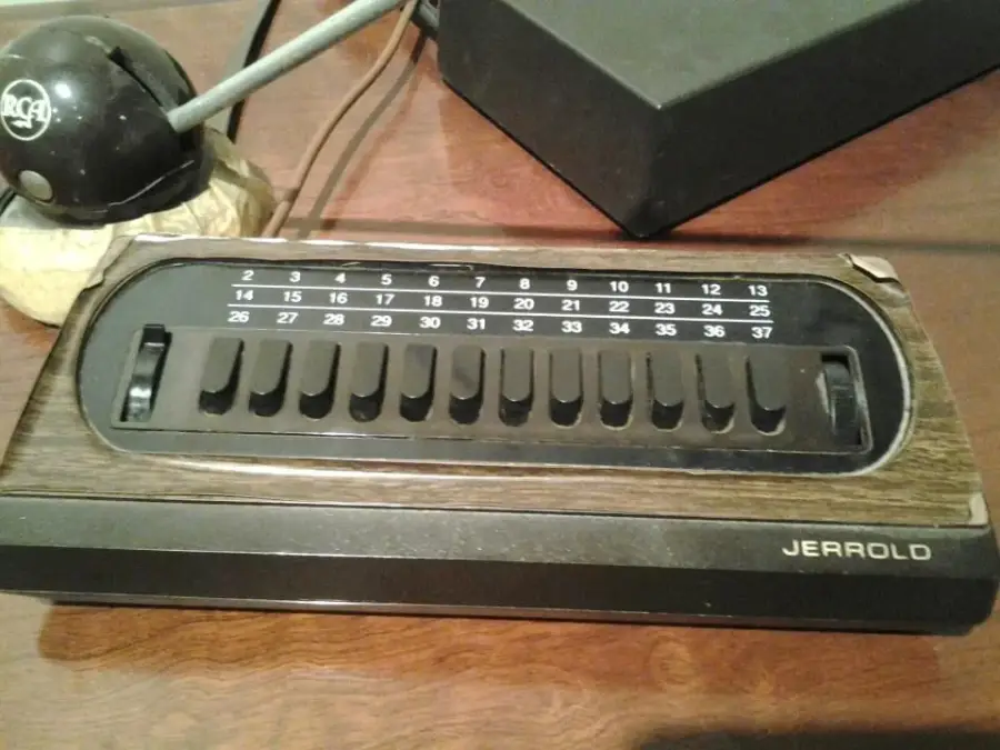 A vintage Jerrold cable TV converter box with a woodgrain finish and 12 black channel selector levers, numbered 2 to 37, sits on a wooden table next to an RCA device.