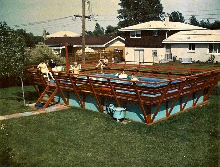 A group of children and adults enjoy an above-ground backyard pool surrounded by a wooden deck, with suburban houses and lawns visible in the background.