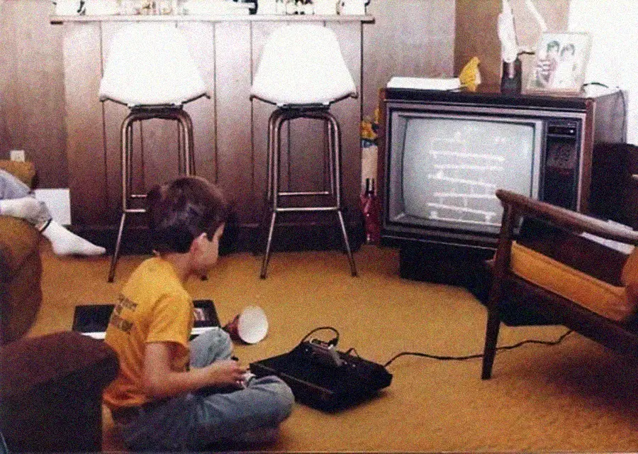 A child sits on a carpeted floor playing a vintage video game console connected to an old CRT television in a cozy, retro living room with two white barstools and wood-paneled walls.