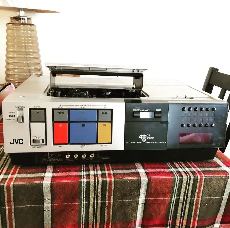 A vintage JVC VHS video cassette recorder sits on a plaid tablecloth, with the tape compartment open and various large buttons and input ports visible on the front panel. A lamp and chair are in the background.