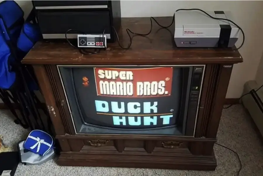 A vintage TV in a wooden cabinet displays the "Super Mario Bros. / Duck Hunt" game title screen, with a classic Nintendo Entertainment System (NES) console and controller on top.