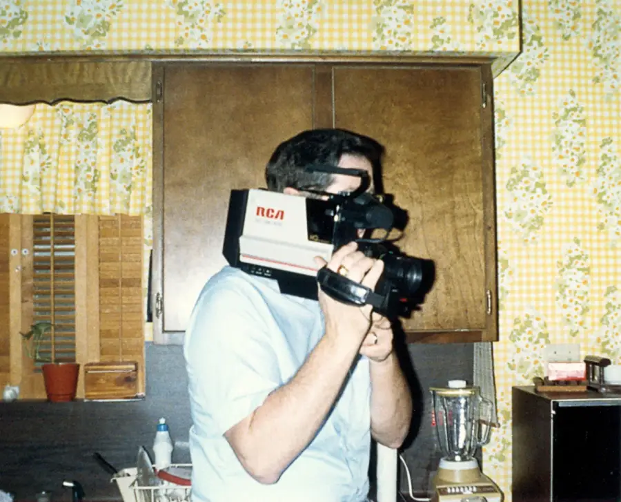 A person in a light blue shirt holds a large vintage RCA video camera on their shoulder in a retro kitchen with wooden cabinets and yellow floral wallpaper. A blender and various kitchen items are on the counter.