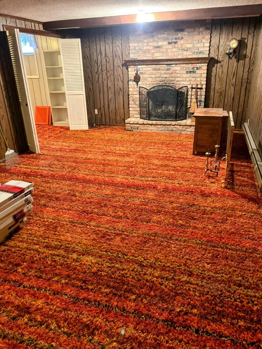 Basement room with orange shag carpet, wood-paneled walls, a brick fireplace with a black screen, built-in shelves, a folding door, and scattered furniture and boxes. Warm lighting creates a retro atmosphere.