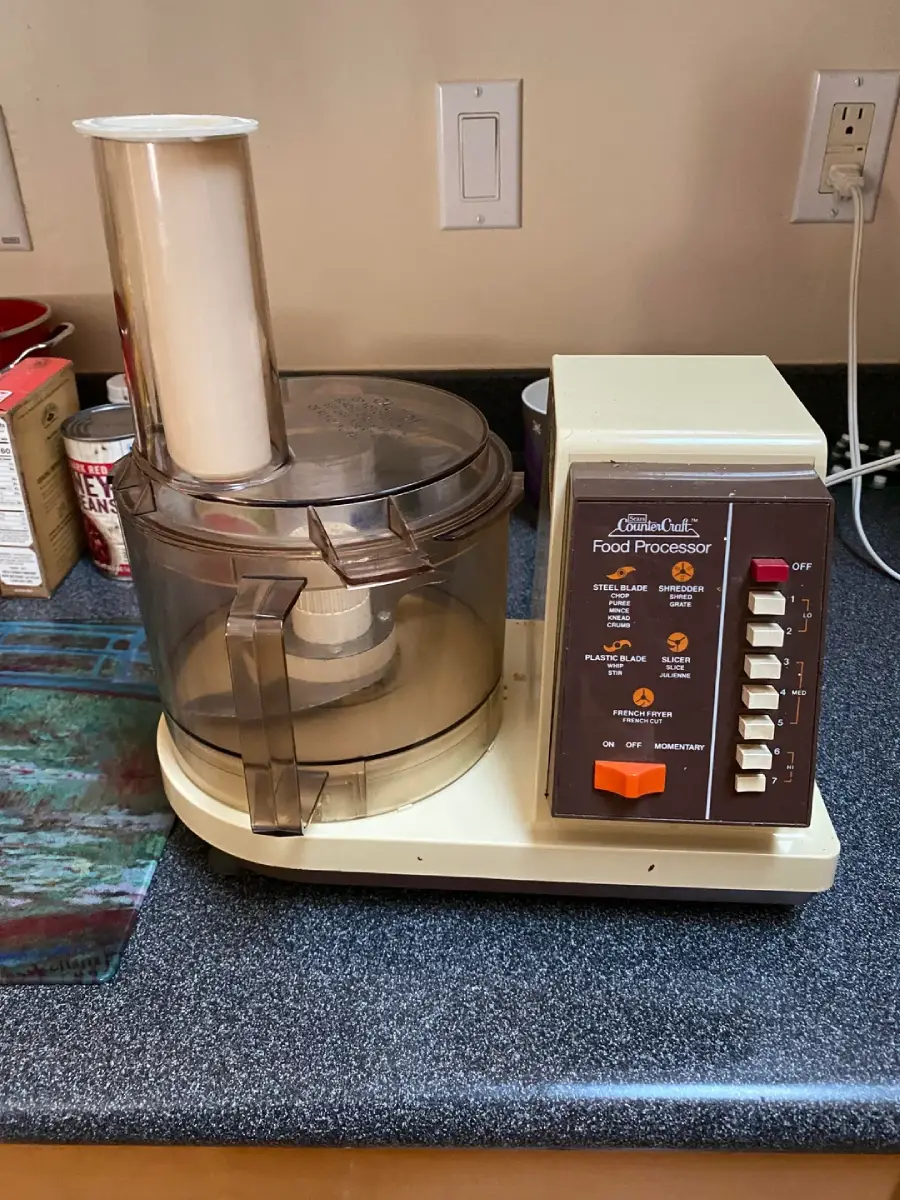A vintage beige and brown food processor with labeled buttons sits on a kitchen counter near a glass cutting board and canned goods. The appliance has a large clear mixing bowl and lid with a tall feed chute.