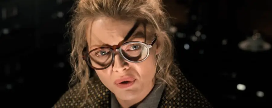 A woman with tousled blonde hair wears large, eccentric glasses with overlapping lenses, looking surprised or concerned in a dimly lit room.