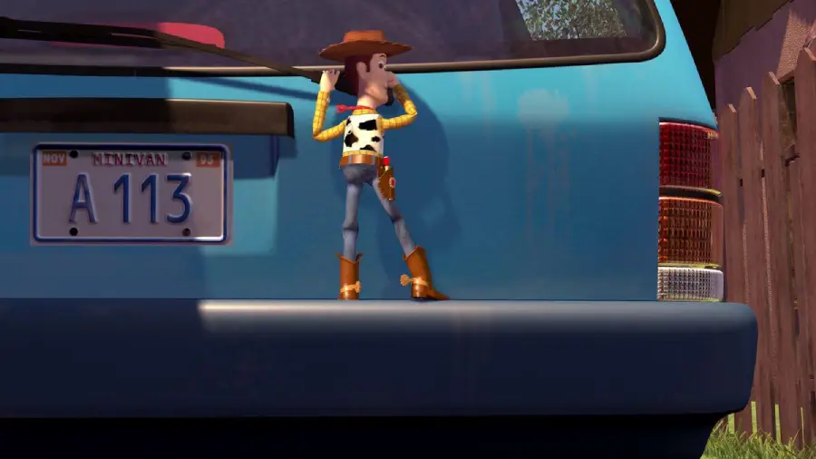 Woody, a cowboy toy wearing a hat and boots, stands sneaking behind the rear of a blue car with a license plate reading "A 113." The scene is outdoors near a fence and some grass.