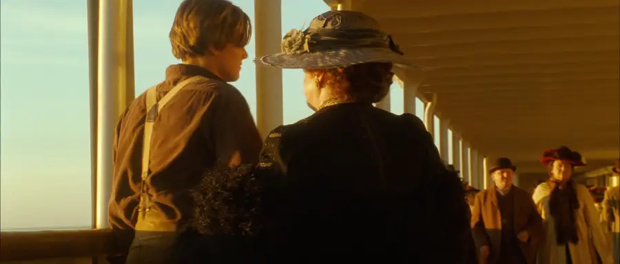 A young man in suspenders and a woman in a decorative hat walk along a sunlit ship deck, with other passengers visible in the background.