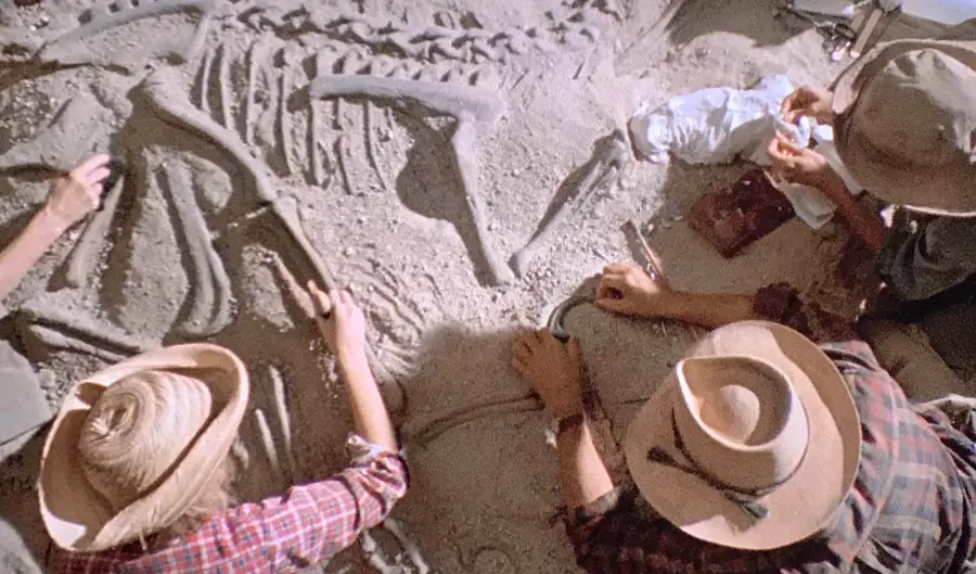 Three people wearing hats and plaid shirts carefully excavate large dinosaur fossils from sandy ground, using small brushes and tools. The bones form part of a skeleton, partially uncovered in the dirt.