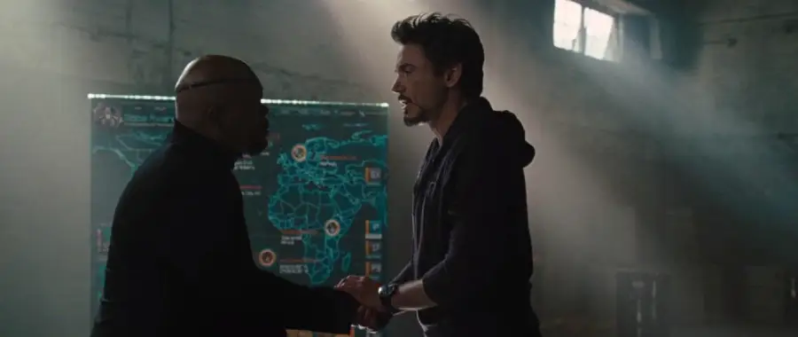 Two men stand facing each other in a dimly lit room, one bald and wearing glasses, the other with dark hair and a beard. A large digital map with bright graphics is visible in the background.