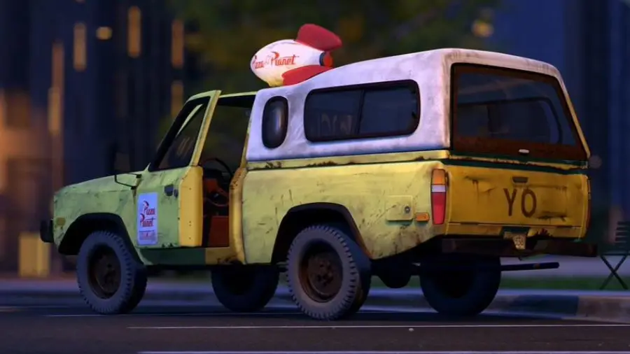 A yellow, weathered pickup truck with a white camper shell and a large rocket on top, displaying the "Pizza Planet" logo, parked on a street at dusk.