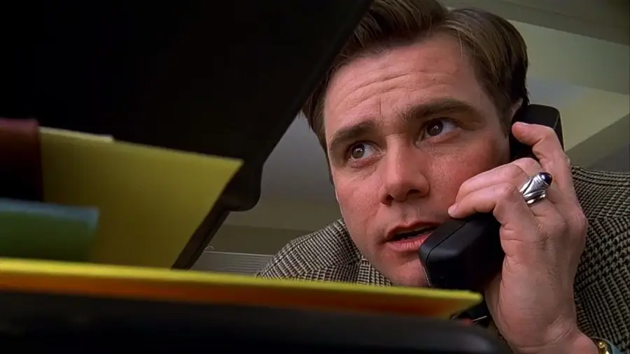 A man with light brown hair, wearing a suit jacket and a silver ring, holds a black phone to his ear while leaning over a desk with colorful folders in the foreground. He appears to be listening intently.