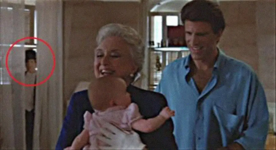 A smiling woman holds a baby while a man stands beside her. In the background, a child partly obscured by a curtain appears circled in red. The scene occurs in a warmly lit room with shelves and decor visible.