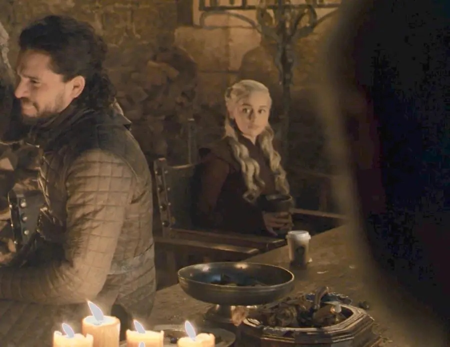 A medieval-style dining scene with a woman with long, braided white hair holding a cup, sitting beside a man in armor. There are candles and food on the table, and a modern coffee cup is visible.