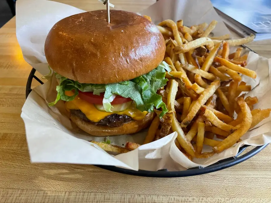 A cheeseburger with lettuce and tomato on a bun is served in a basket with a generous portion of thin, crispy French fries on parchment paper, set on a wooden table.