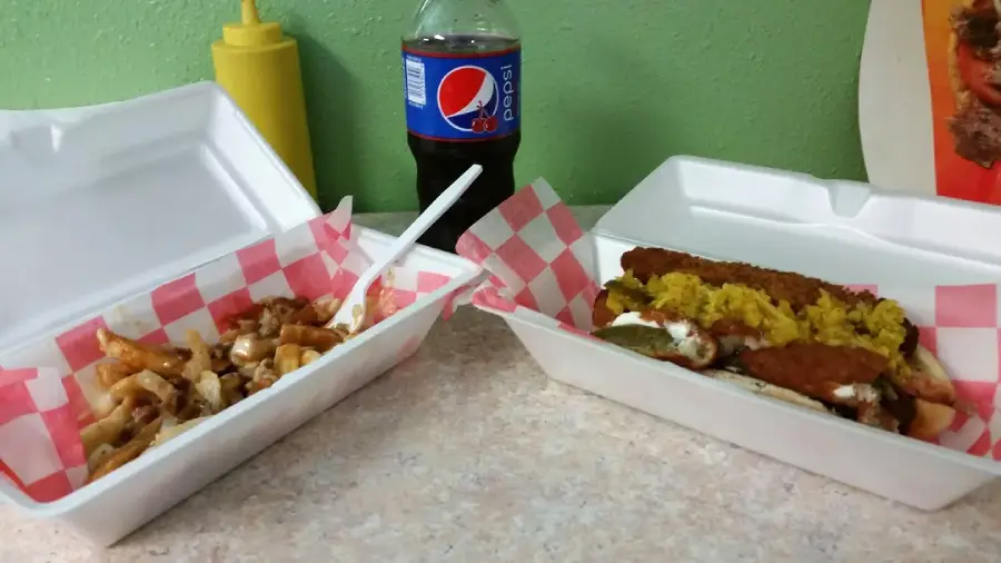Two open takeout containers on a table: one with fries topped with sauce and a plastic fork, the other with yellow rice, breaded chicken strips, and sauce. A bottle of Pepsi and a yellow squeeze bottle are in the background.