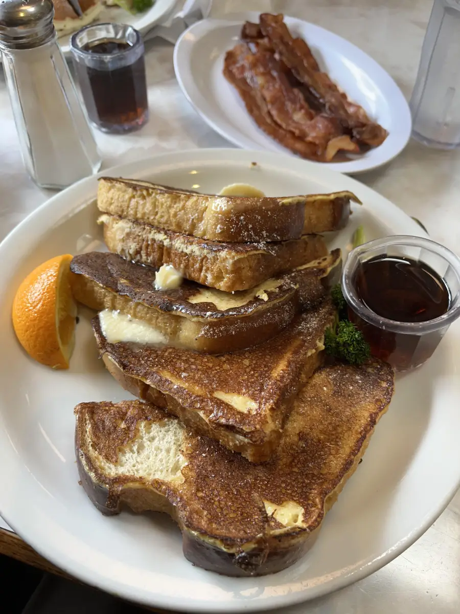 A plate of French toast slices topped with butter, served with a slice of orange, parsley, and a cup of syrup. In the background, a plate of bacon and a glass of water are visible.