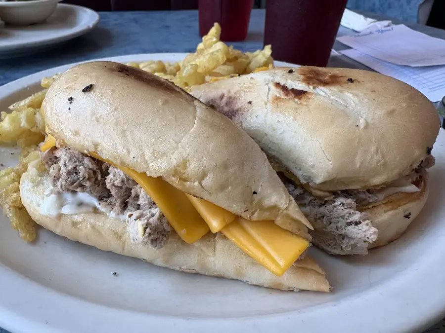 Two sub sandwiches on a white plate, filled with shredded meat, cheddar cheese slices, and mayonnaise, served with a side of crinkle-cut fries. Drinks and papers are visible in the background.