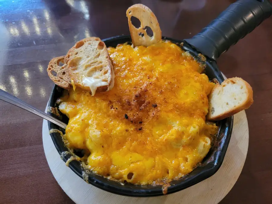 A skillet filled with baked macaroni and cheese, topped with melted cheddar and served with two slices of toasted baguette, sits on a wooden coaster. A spoon rests in the dish.
