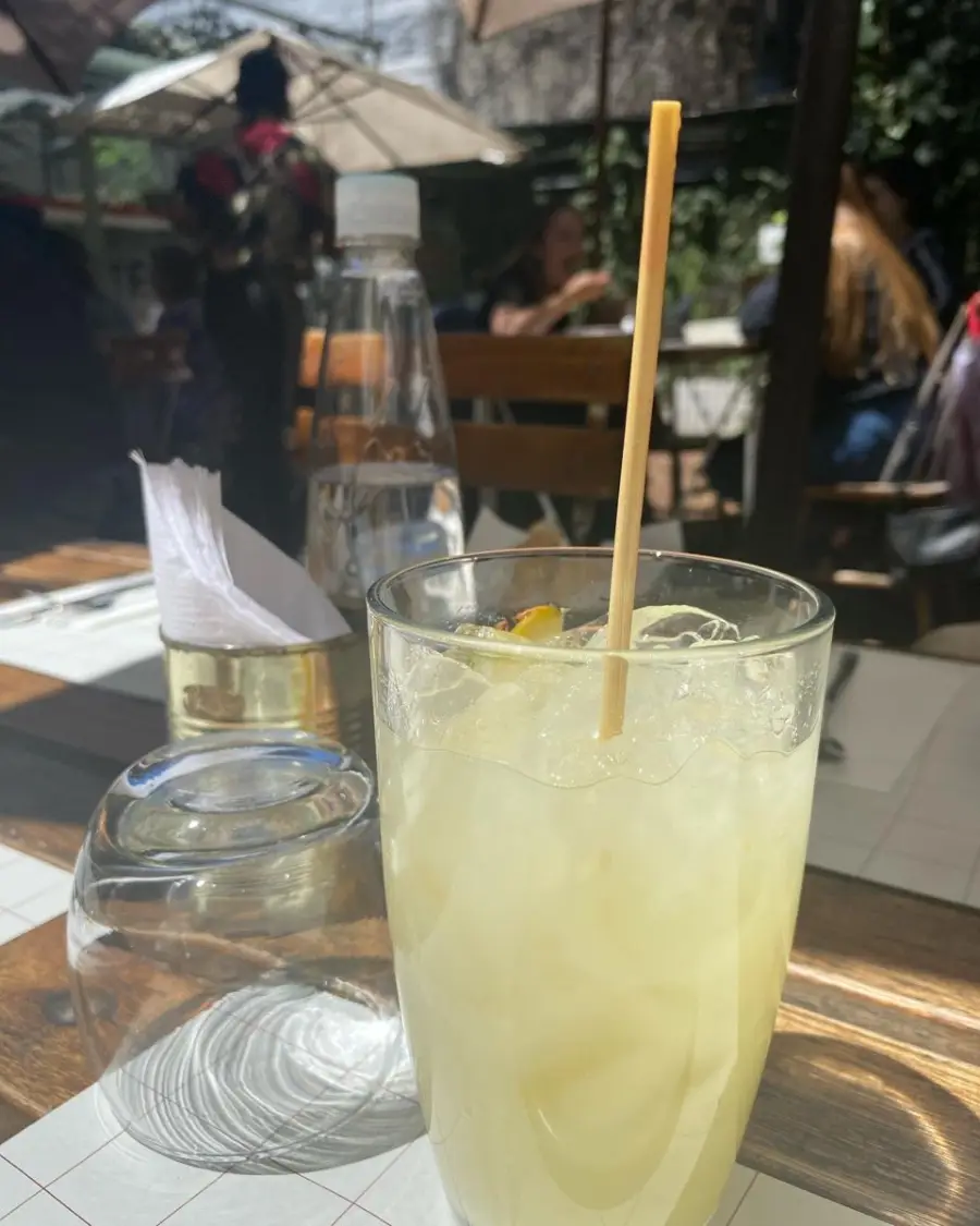 A glass of lemonade with ice and a straw sits on an outdoor table with sunlight casting shadows. A glass, a bottle of water, napkins, and blurred people in the background complete the scene.