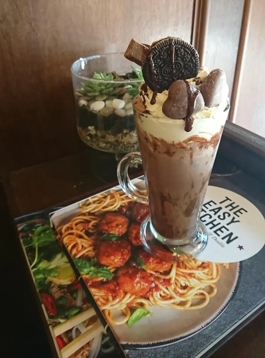 A tall glass of chocolate milkshake topped with whipped cream, an Oreo cookie, chocolate drizzle, and chocolates, placed on a cookbook with a spaghetti dish on the cover, next to a small decorative plant.