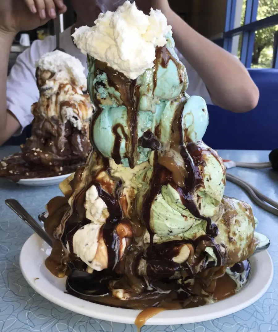 A large ice cream sundae with scoops of mint, vanilla, and blue ice cream, topped with whipped cream, chocolate sauce, and caramel sauce, sits on a white plate. Another sundae is visible in the background.