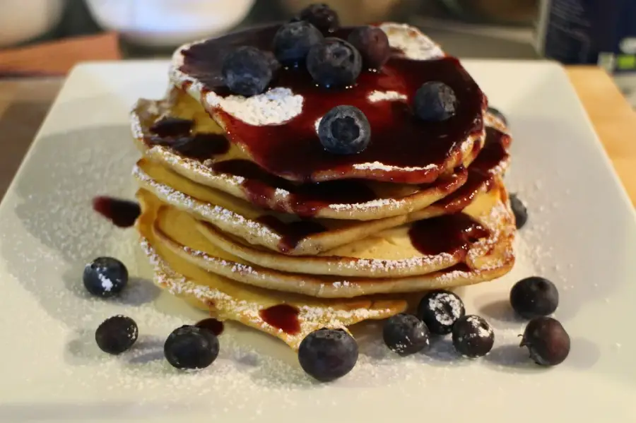 A stack of pancakes topped with blueberry sauce and fresh blueberries, dusted with powdered sugar, served on a white plate. More blueberries are scattered around the plate.