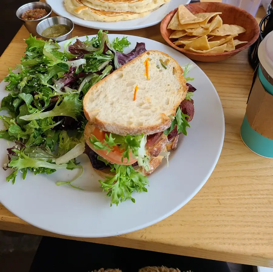 A plate with a sandwich filled with lettuce, tomato, and bacon, served alongside a mixed greens salad. Tortilla chips, sauces, and a cup with a cardboard sleeve are on the table.