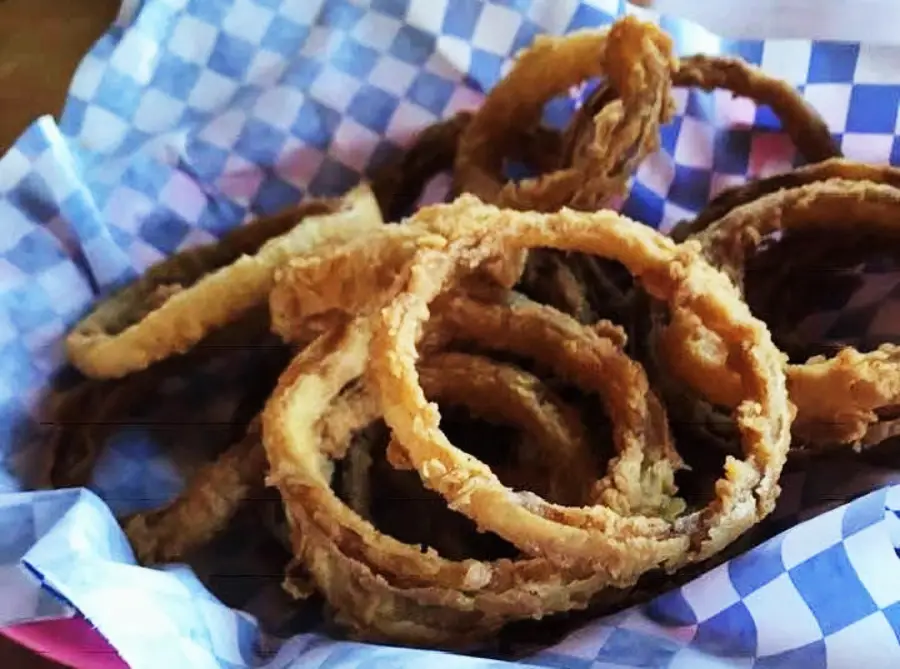 A close-up of crispy, golden-brown onion rings served in a basket lined with blue and white checkered paper.