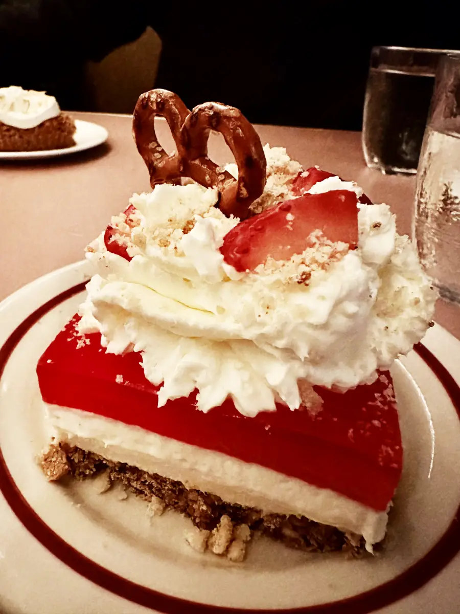 A slice of layered dessert with a crumbly crust, creamy middle, and red gelatin top, topped with whipped cream, fresh strawberries, and two pretzels, served on a white plate with red trim.